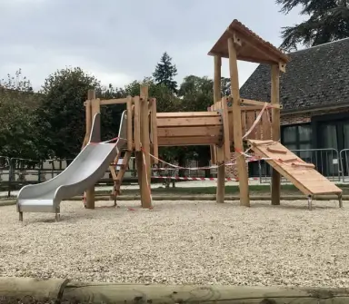 La nouvelle aire de jeux terminée sur la place du village : structure de jeu en bois avec toboggan métallique, devant l'école.