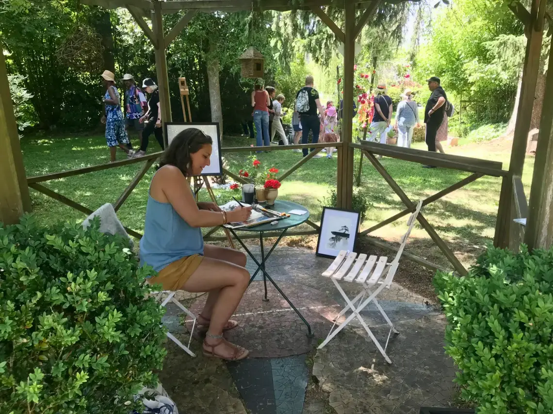 Une artiste peint à l’aquarelle dans le jardin lors de la Fête de la Peinture, ses œuvres exposées et des visiteurs en arrière-plan.