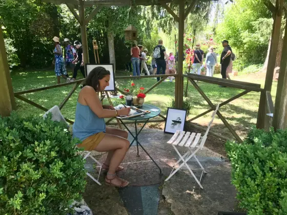 Une artiste peint à l’aquarelle dans le jardin lors de la Fête de la Peinture, ses œuvres exposées et des visiteurs en arrière-plan.