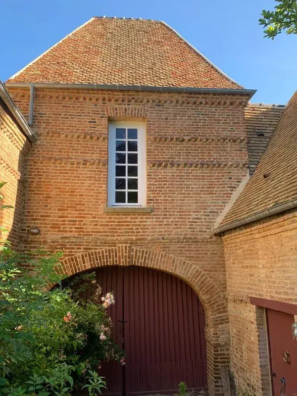 Pignon et porche en brique aux portes rouges, encadrés de rosiers grimpants.